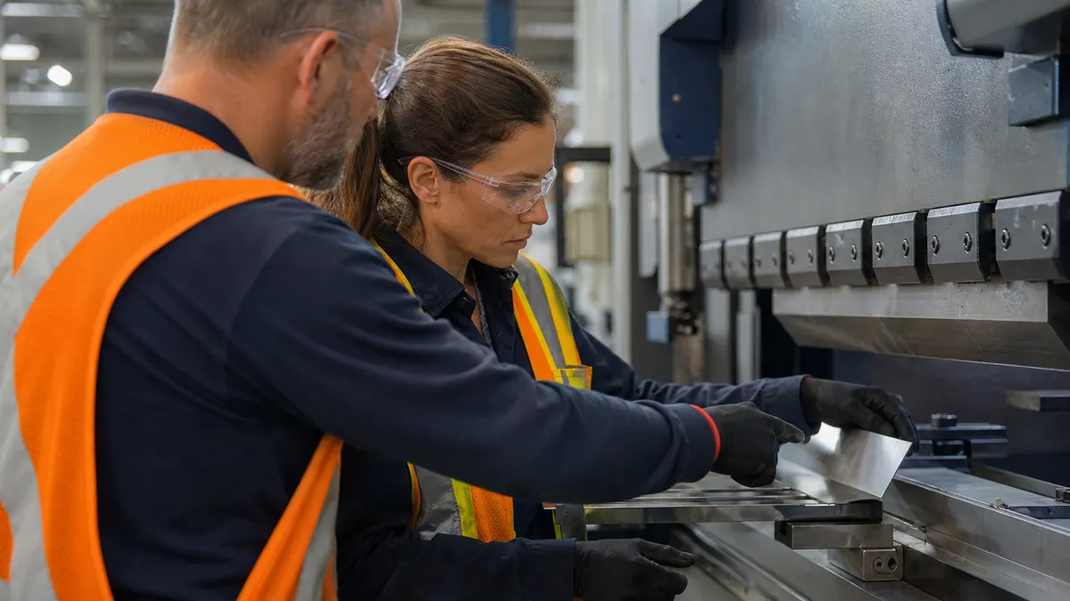 an operator being shown a work instruction at a press brake by a supervisor, both in safety glasses and high-visibility vests